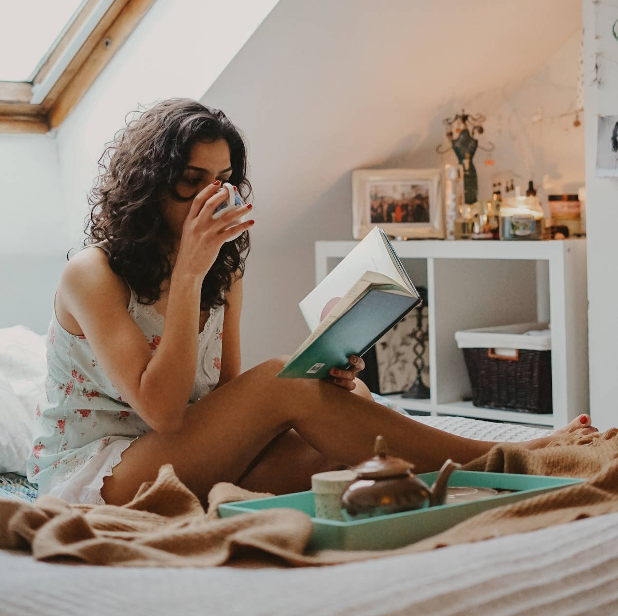 Frau sitzt im Bett, trinkt aus einer Tasse und liest ein Buch